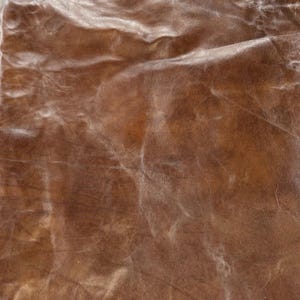 Puede incluir: Primer plano de un trozo de cuero marrón con aspecto envejecido. El cuero tiene un tono rico y cálido con variaciones de color y textura, mostrando pliegues y sutiles reflejos.