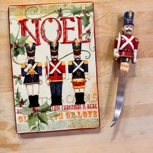 Puede incluir: Una fuente rectangular navideña con la palabra "NOEL" en rojo y tres figuras de soldados de juguete. Un cuchillo de queso decorativo con un mango de soldado de juguete está al lado de la fuente.