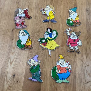 May include: A set of eight flat wooden Christmas ornaments featuring Snow White and the seven dwarfs. Each character is depicted in colourful clothing with holiday accents like candy canes, wreaths, and a stocking labelled with the name Sneezy.