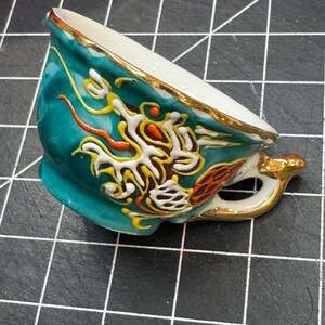 May include: A teal ceramic teacup with a gold-trimmed rim and handle. The cup features a white, yellow, and orange dragon design. The handle and rim are gold. The teacup is on a black and white grid surface.