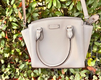 May include: A light gray leather handbag with a top handle and a shoulder strap. The bag has a rectangular shape with a curved top and a small metal logo. The background is a lush green bush.