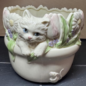 Könnte beinhalten: Cremefarbener Keramik-Blumentopf mit einer weißen Katze mit blauen Augen, umgeben von pastellfarbenen Blumen. Der Topf zeigt Gänseblümchen, Tulpen und kleine lila Blumen und schafft ein charmantes Frühlingsdesign.