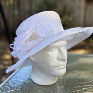 May include: A white wide-brimmed hat with a structured crown and a textured straw-like finish. The hat is adorned with a large, layered white fabric flower accent on the side, featuring delicate ribbon loops and a soft, ruffled petal design.