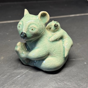 May include: A celadon-glazed ceramic figurine of a koala with a baby koala clinging to its back. The figurine has a crackle glaze and is light green with dark accents on the nose and eyes. The koala is sitting down.