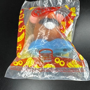 May include: A Mr. Potato Head toy in a clear plastic bag. The toy has a brown head, pink ears, a black mustache, and a blue body. The bag has red and yellow graphics and the Burger King logo.