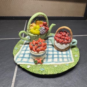 Könnte beinhalten: Ein dekoratives Keramik-Teeservice mit Obstmotiven. Das Set umfasst eine Teekanne in Form eines Korbes mit verschiedenen Früchten, eine Teekanne in Form eines Korbes mit roten Äpfeln und eine Schale mit Erdbeeren, alles auf einem Tablett.