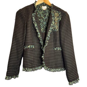 May include: A dark brown textured blazer featuring a ruffled light blue and green patterned trim along the front opening, hem, and cuffs. The jacket includes two front pockets with matching patterned flaps and an embroidered floral detail.