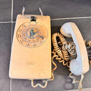 May include: Vintage cream-colored wall phone with a rotary dial and a matching handset. The phone has a metal cradle and a coiled cord. The dial features numbers and letters. The phone is a Bell System model.
