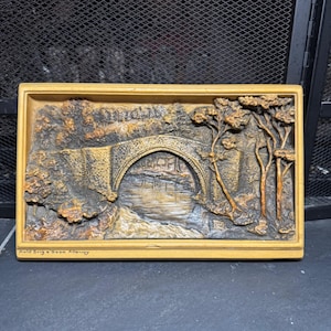 May include: A rectangular wall plaque featuring a relief sculpture of a stone bridge over a river, surrounded by trees. The scene is rendered in shades of brown and tan. The text Auld Brig o Doon Alloway is printed in the bottom left corner.