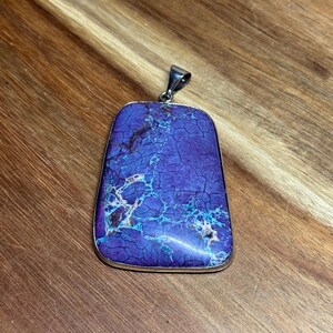 May include: A rectangular pendant featuring a vibrant purple stone with a complex network of dark brown and bright turquoise veining. The stone is set in a smooth silver-toned metal frame with a small, polished silver-toned bail at the top.