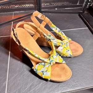 Puede incluir: Sandalias de cuña con una correa de cuero marrón claro y un lazo de tela estampada con limones. Las sandalias tienen un tacón de cuña de corcho y una plantilla de ante marrón claro.