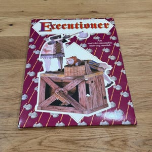 May include: A rectangular box for an easy-to-assemble moving model titled Executioner. The packaging features a deep magenta background with a repeating pattern of silver axes and a central illustration of a figure standing by a wooden structure.