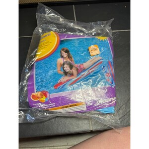 Op de afbeelding: Een opblaasbaar zwembadsurfboard in een doorzichtige plastic verpakking. De productverpakking toont een afbeelding van een persoon die een rood, wit en blauw surfboard in een zwembad gebruikt. De tekst op de verpakking bevat het woord Surfboard en de afmetingen 114 cm bij 48 cm bij 13 cm.