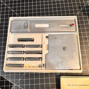 Puede incluir: Un kit de ojal vintage en una caja de plástico blanco. El kit incluye varios componentes metálicos, incluyendo sellos, un mecanismo deslizante y una pequeña placa de metal. La caja está abierta, revelando las herramientas en el interior. El fondo es una rejilla negra.