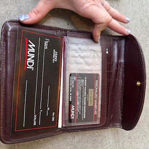 Könnte beinhalten: Eine bordeauxfarbene Lederbrieftasche, geöffnet, um Kartenfächer und Einsätze freizugeben. Die Brieftasche trägt das "Mundi"-Logo und den Text sowie eine "Personalized Nameplate"-Karte. Die Brieftasche wird von einer Person offen gehalten.