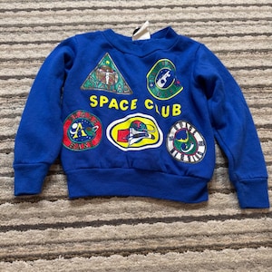 Può includere: Una felpa blu reale a maniche lunghe con scollo rotondo e polsini a coste. La parte anteriore presenta la scritta gialla SPACE CLUB circondata da cinque toppe ricamate colorate che raffigurano vari grafici a tema spaziale, inclusi razzi e pianeti.