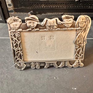 May include: A silver-colored picture frame with a rectangular opening. The frame is decorated with relief carvings of five children's faces across the top and feet at the bottom. The sides feature floral and vine designs.