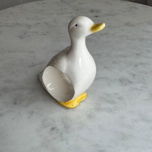 May include: A ceramic duck figurine with a white body, yellow beak, and yellow feet. The duck is standing on a marble surface. The figurine has a classic, simple design.