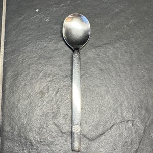 Könnte beinhalten: Ein Edelstahl-Löffel mit einer abgerundeten Schüssel und einem langen, schlanken Griff. Der Griff hat ein strukturiertes Design und ein kleines, kreisförmiges Emblem am Ende. Der Löffel ist zum Essen oder Servieren von Speisen bestimmt.