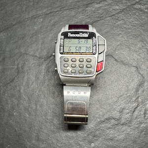 May include: A silver Famous Trails calculator watch with a digital display showing the date and time. The watch has a calculator keypad and a red power button. The watch band is silver and has a clasp.