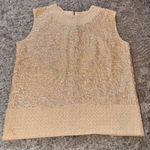 May include: A sleeveless, cream-colored top with a textured, sequined design. The top features a round neckline and a zipper closure at the back. The bottom hem has a detailed, beaded border, creating a vintage aesthetic.