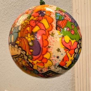 Könnte beinhalten: Ein farbenfroher, runder Ornament mit psychedelischem Design. Das Ornament zeigt eine lebendige Mischung aus orangefarbenen, rosa, gelben, grünen und blauen Mustern. Es wird von einer grünen Schnur gehalten, was darauf hindeutet, dass es zur Dekoration dient, möglicherweise als Weihnachtsbaumschmuck.