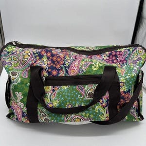 May include: A colorful paisley-patterned duffel bag with a green background and accents of pink, purple, and yellow. The bag has a brown zipper and black handles. The bag is designed for travel or carrying items.