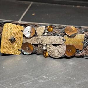 May include: A braided belt made of dark brown and black leather strands, featuring a decorative buckle assembly with a rectangular gold-toned metal plate, four round brown buttons, a silver-toned cross-shaped metal piece, and amber beads.