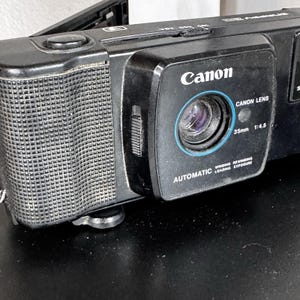 May include: Black Canon camera with a 35mm 1:4.5 lens. The camera has the words "Canon Lens", "Automatic Winding Rewinding Loading Exposure" printed on the front. The camera has a textured grip on the side.