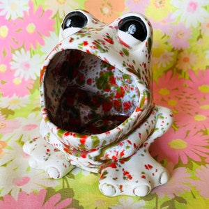May include: A white ceramic frog planter with red, green, and white speckled glaze. The frog has a wide open mouth and large, black eyes.