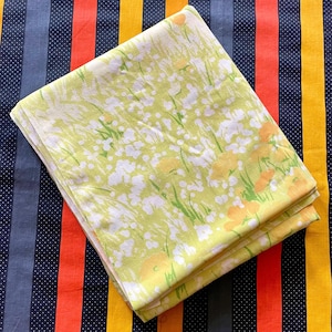 May include: A folded piece of fabric featuring a light green background with a white and yellow floral pattern. The print consists of abstract white flower clusters and larger yellow blossoms scattered across the light green surface.