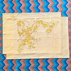 May include: Two cream-colored pillowcases with a floral design featuring yellow and pink roses and green leaves. The pillowcases are rectangular and have a decorative border. The background is a blue, pink, and brown patterned blanket.