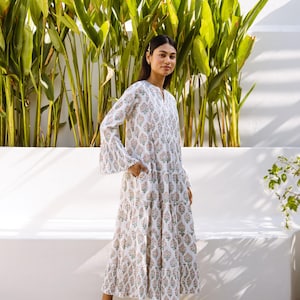 May include: A white maxi dress with a floral print and bell sleeves. The dress has a tiered skirt and is worn by a person standing in front of a white wall and green plants.