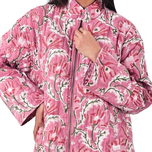 May include: A pink quilted jacket with a floral pattern in red, white, and green. The jacket has a stand-up collar, long sleeves, and a full-length zipper. The floral design is repeated across the entire jacket.