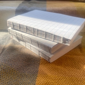 May include: Three white notebooks stacked on top of each other. The top notebook has lined pages and a white cover. The bottom two notebooks have white covers.