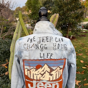 May include: Light blue denim jacket with the text "ONE JEEP CAN CHANGE YOUR LIFE" in black script. The back features a mountain and sun design with the word "Jeep" in a red and white font.