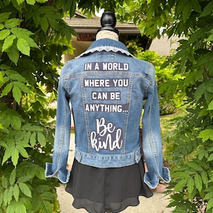 May include: A blue denim jacket with a white lace collar and patches on the back that say "In a world where you can be anything... Be kind".