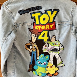 Può includere: Giacca di jeans azzurra con una grande grafica di "Toy Story 4" sul retro. La grafica include Woody, Buzz Lightyear e altri personaggi. È visibile anche il logo Disney Pixar.