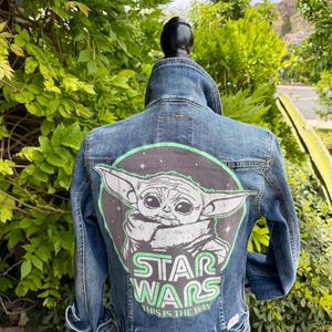 May include: A blue denim jacket featuring a graphic of Grogu from Star Wars on the back. The design includes the words "STAR WARS" in green and the phrase "THIS IS THE WAY". The jacket has a classic denim style.