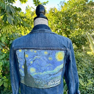 Pode incluir: Jaqueta jeans azul com um patch colorido retratando a pintura "Noite Estrelada" de Van Gogh nas costas. O patch inclui o texto "BEYOND Van Gogh". A jaqueta tem botões e mangas dobradas.