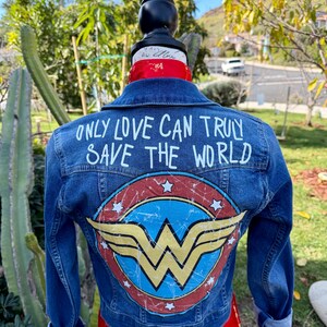May include: A blue denim jacket with a Wonder Woman logo on the back. The logo is a circular design with a red background, white stars, and a gold emblem. The text "ONLY LOVE CAN TRULY SAVE THE WORLD" is written above the logo in white.