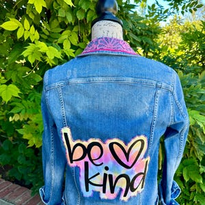 Może przedstawiać: Jasnoniebieska kurtka dżinsowa z efektem zniszczenia. Z tyłu kurtki znajduje się kolorowa naszywka tie-dye z napisem "be kind" i sercem. Kołnierz ma różowo-białe wzorzyste wykończenie.