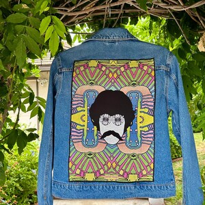 May include: A light blue denim jacket with a colorful graphic on the back. The graphic features a black and white face with a mustache and glasses, set against a vibrant, geometric background with pink, green, and yellow accents.