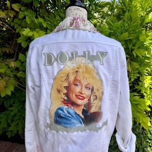 Może przedstawiać: Biała kurtka dżinsowa z portretem Dolly Parton i napisem "DOLLY". Kurtka ma kwiatowy kołnierz i jest prezentowana na manekinie. Tło to zielone liście.