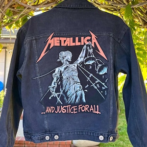 May include: Dark grey denim jacket with a large patch featuring the band name "Metallica" in orange and a graphic of Lady Justice. The text "...AND JUSTICE FOR ALL" is below the graphic.