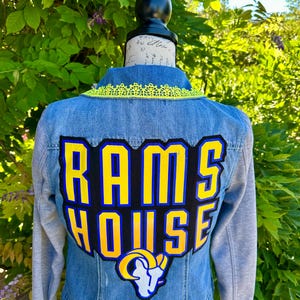 Pode incluir: Casaco de ganga com mangas cinzentas e gola de renda amarela. As costas apresentam um grande patch com as palavras "RAMS HOUSE" em amarelo e azul, juntamente com um logótipo da equipa. O casaco é exibido num manequim.