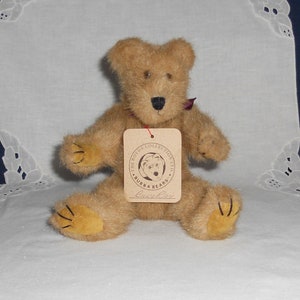 Puede incluir: Un oso de peluche de color marrón claro con una nariz negra y una pequeña cinta roja. El oso tiene una etiqueta que dice "The Boyds Collection Ltd. Bubba Bears" y "Buzby Roy". El oso está sentado con las piernas extendidas.