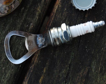 May include: A metal bottle opener with a spark plug attached to it. The opener is rusty and the spark plug is silver with a white ceramic insulator. The spark plug is labeled "NGK  R  1871".