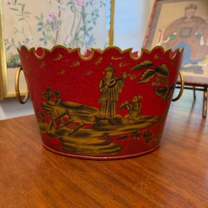 May include: A red oval metal bucket with a scalloped top edge and two gold-toned side handles. The exterior features a black and gold painted scene of figures playing musical instruments, foliage, and rocks, finished with a visible crackle texture.