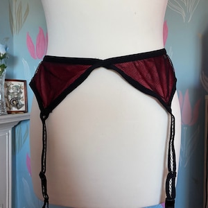 May include: A red mesh garter belt with black edging and adjustable straps. The garment is displayed on a white mannequin. The background features floral wallpaper.